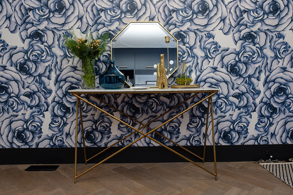 Bold blue rose wallpaper covers a wall with a brass entry table set with a hexagon mirror, gold giraffe and a blue vase