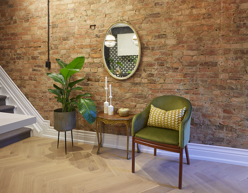 A green chair, console table and palm plant sit in front of a large red brick wall