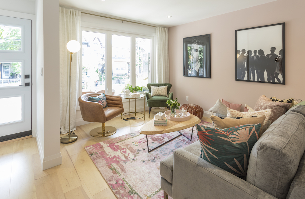 Fully renovated living room with triangular wooden coffee table, pink patterned rug, grey velvet couch and three large windows