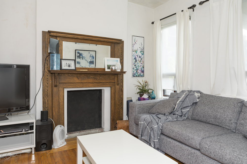 Old fireplace and wood mantel in a messy living room with a grey couch, white coffee table and TV