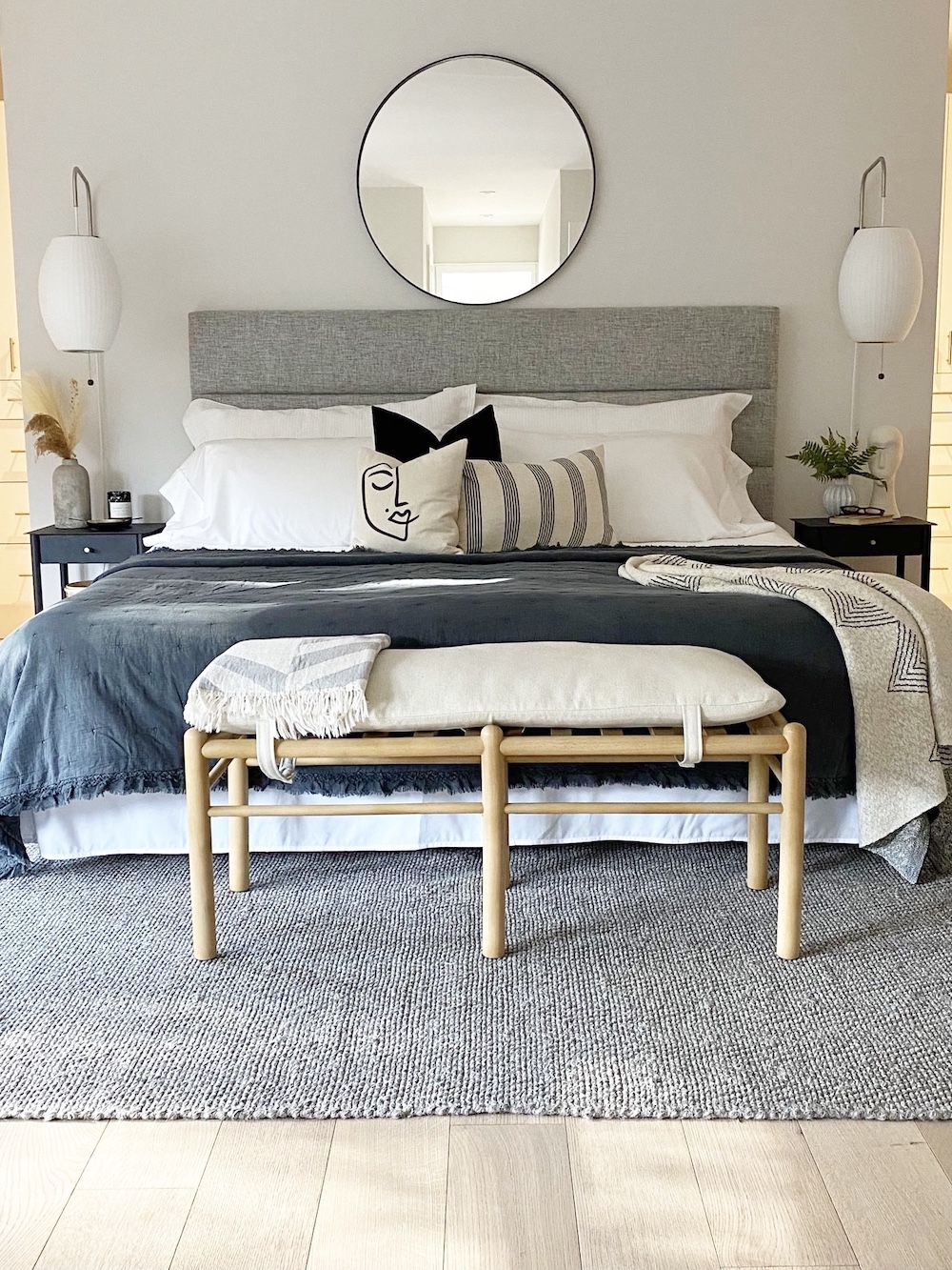 Calm bedroom designed and styled by The Property Stylist Inc. with a large bed with a large upholstered grey headboard, white pillows, a blue throw, a bench sitting at the end of the bed, two large wall sconces on either side of the bed, and large round mirror above the bed