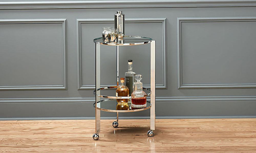 Small round chrome bar cart on wheels with two glass shelves holding glasses, a cocktail shaker and liquor bottles