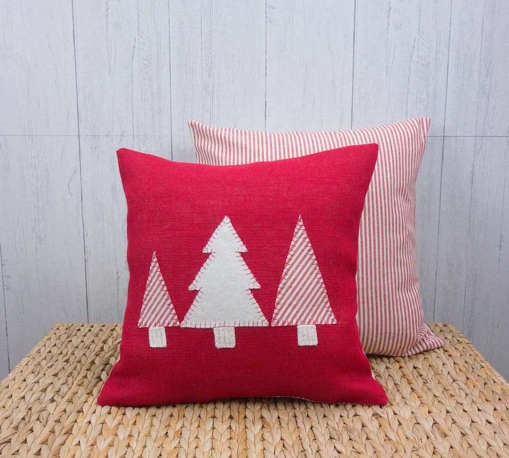 Two red and white holiday themed pillows with applique trees and stripes sit on a grass bench in front of a grey wood plank wall