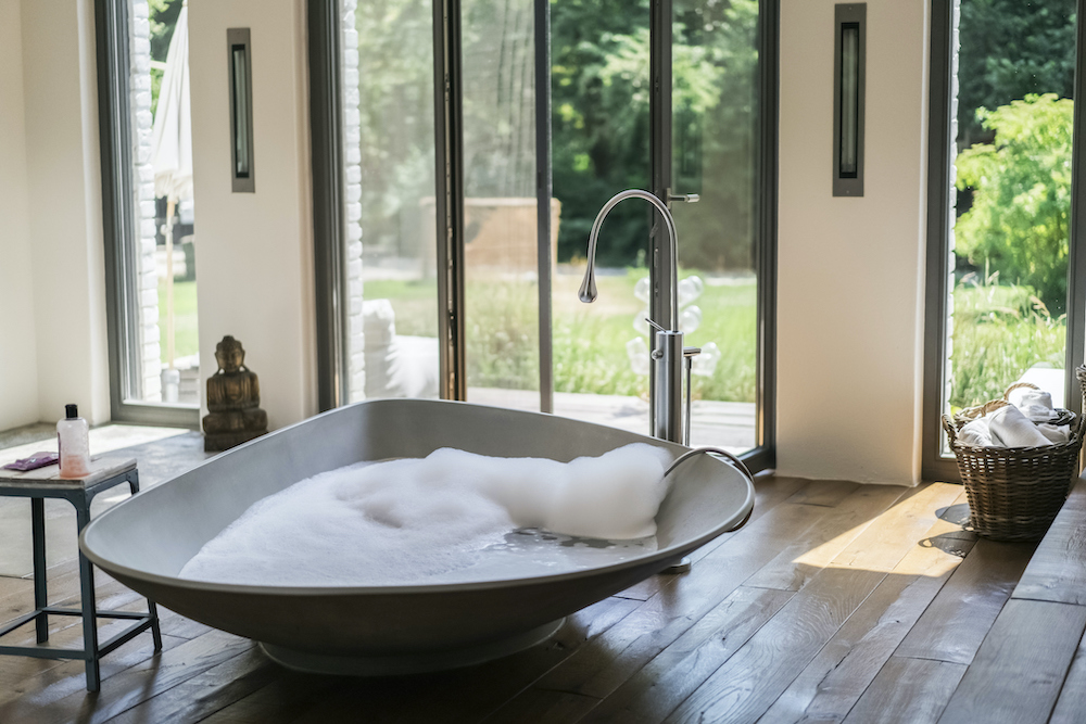Beautiful bathroom with dark wide plank wood floors, a triangular standalone bathtub filled with bubbles, a stoop with a bubble bath bottle and facecloth, large floor to ceiling glass doors going out into a yard, a buddha statue and a basket filled with rolled towels