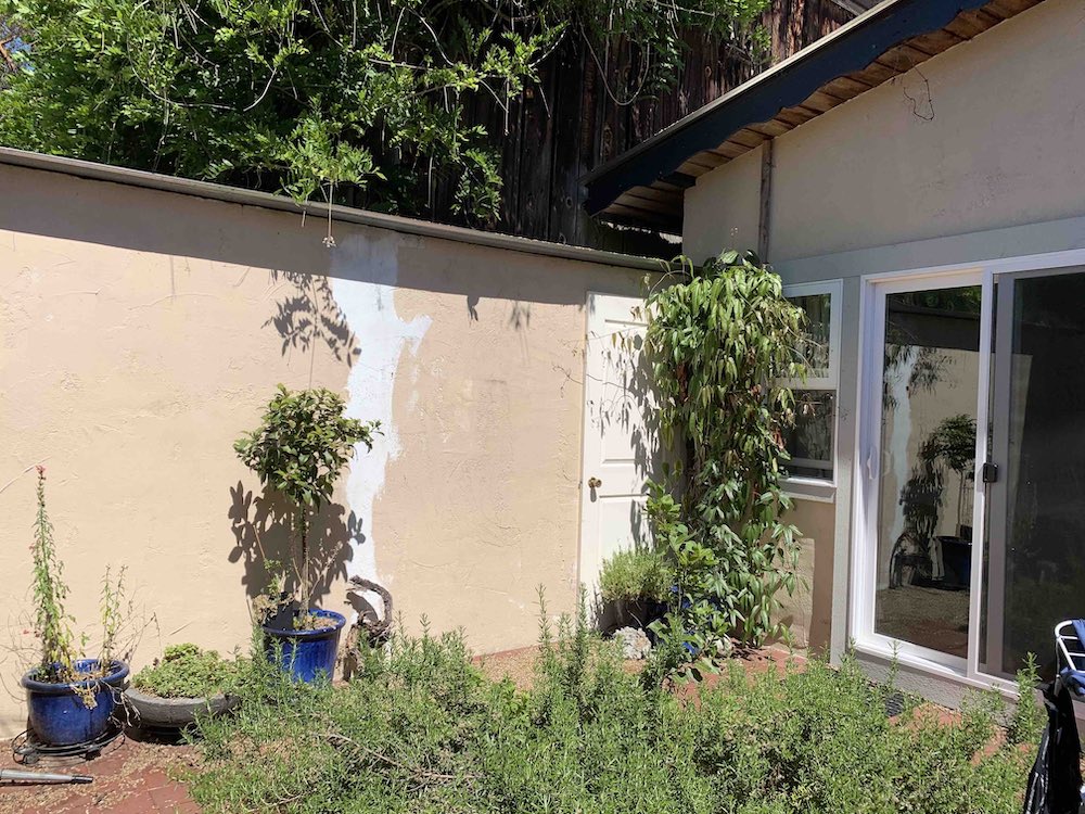 Exterior of a rundown cottage courtyard painted in a beige colour and filled with overgrown plants in pots.