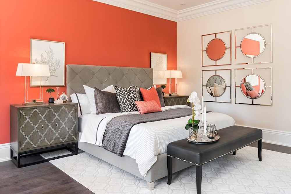 Luxurious guest room with a bold orange coloured feature wall with two pieces of framed artwork behind a bed with a large grey tufted headboard, white sheets and a stack of grey and orange throw pillow, two grey nightstands, and four decorative mirrors on the wall beside the bed