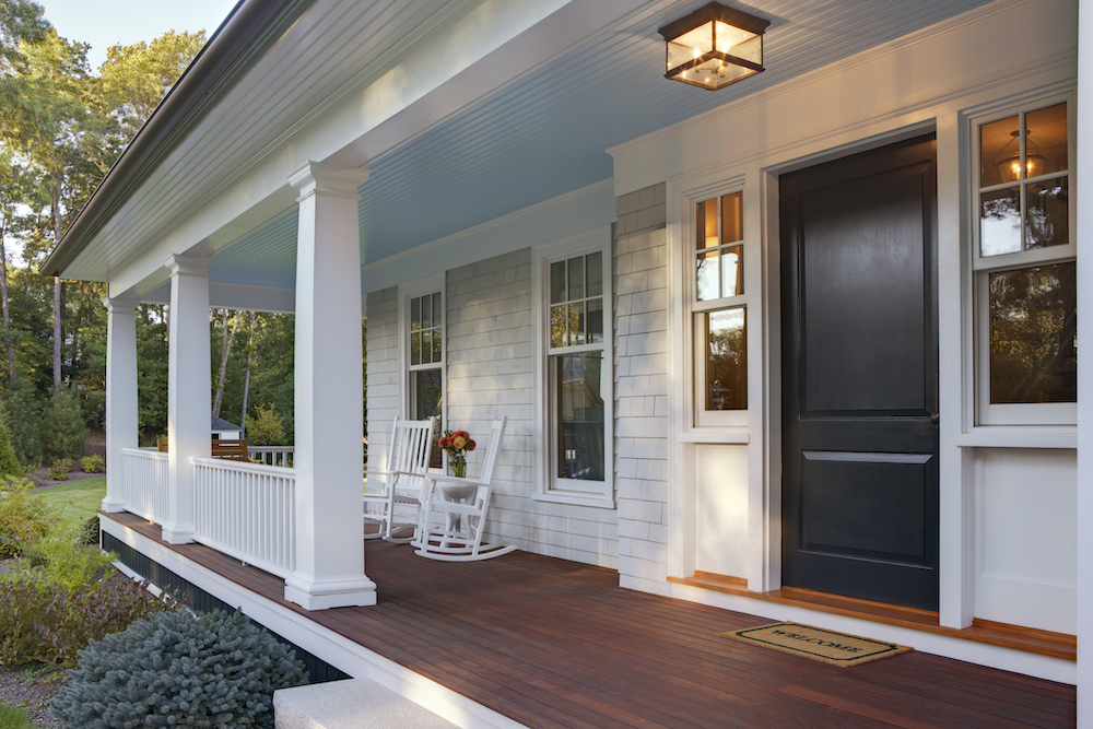Front porch of a beautiful craftsman house with light grey shingles, a blue porch ceiling, a dark blue door with a core welcome mat, dark stained wooden porch floor, and modern light fixture in front of the door