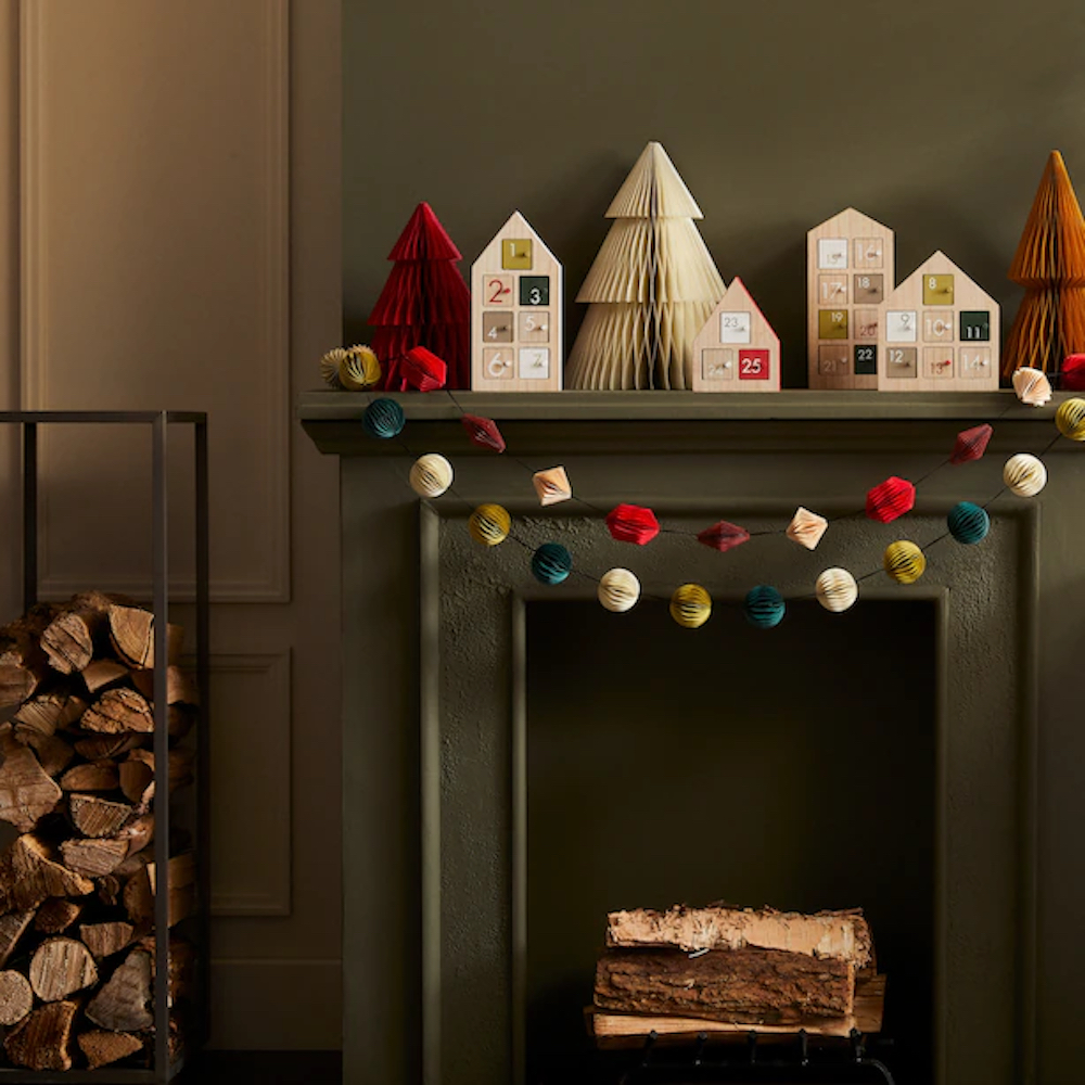 Oui geometric paper garlands hang from green mantle as part of a Christmas decoration display with Oui accordion paper trees and wooden advent calendar houses