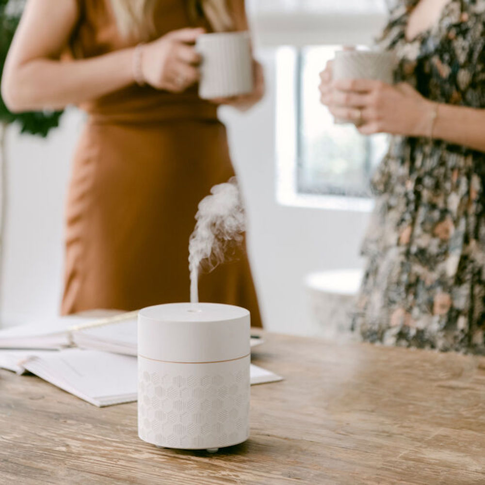 A white textural Saje diffuser, designed with The Little Market’s co-founders supports a mission to break the cycle of poverty for artisans in marginalized communities, sits on a wood countertop with two women holding cups in the background