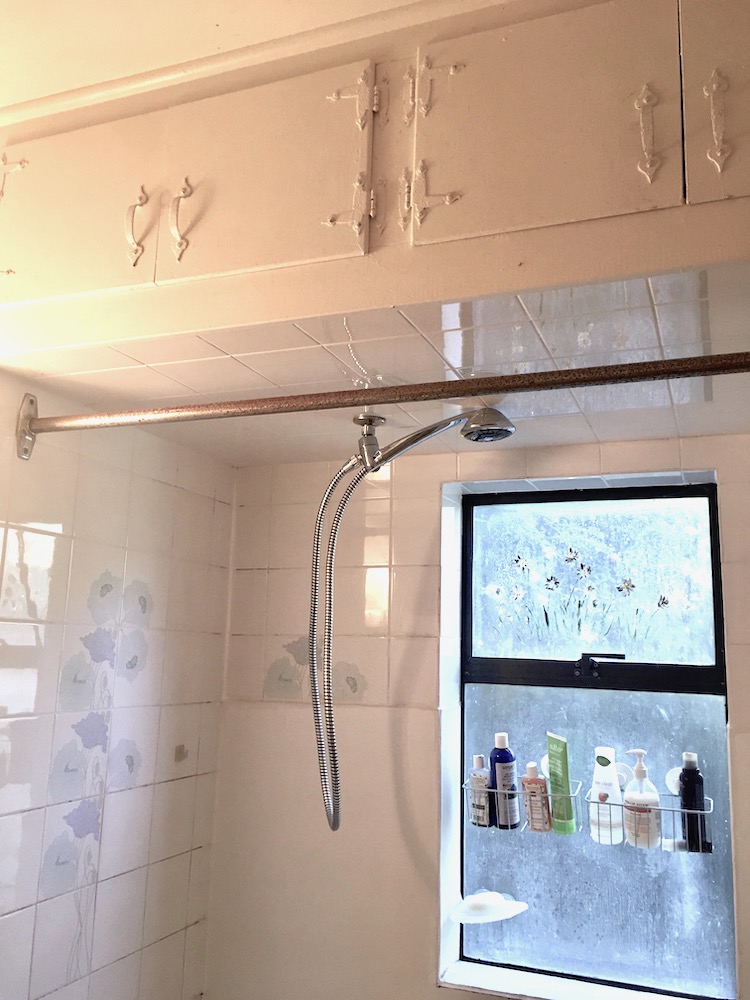 An old bathroom from the 1980s with a rusted shower rail, a window with product baskets, and a removable shower handle