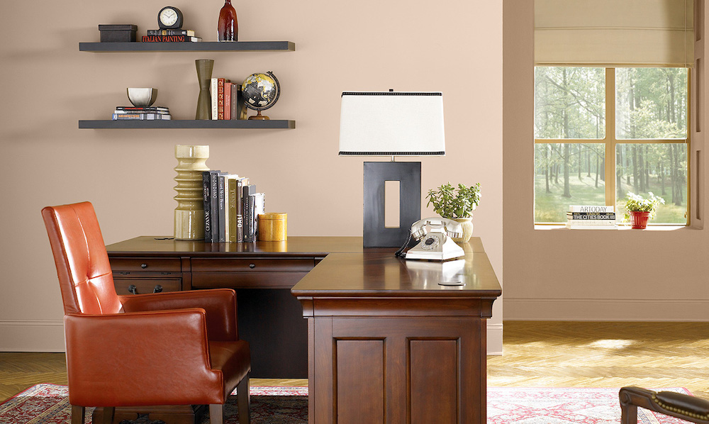 Masculine office with large L-shaped desk, orange leather chair, two brown floating shelves with books and art objects, and walls painted in BEHR Venetian Mask PPU3-06 and Soft Chamois PPU4-04