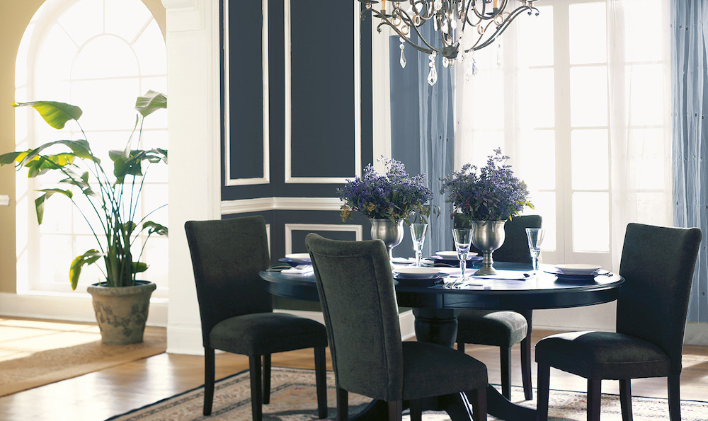 Traditional French style dining room with a round blue dining table set for a meal, two silver vases with purple flowers, four dark grey upholstered dining chairs, a crystal chandelier, a large palm plant, a large window with blue drapes, and walls and trim painted with BEHR Midnight Blue N480-7, Polar Bear 75 and Perennial Gold S310-4