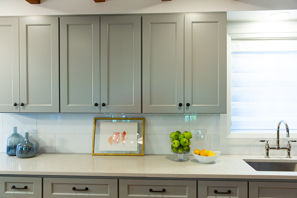 Beautiful putty-coloured cabinets in a new kitchen with a white tile backsplash, cream coloured quarts countertops with a gold framed piece of art, a bowl of apples and a bowl of lemons sitting on it