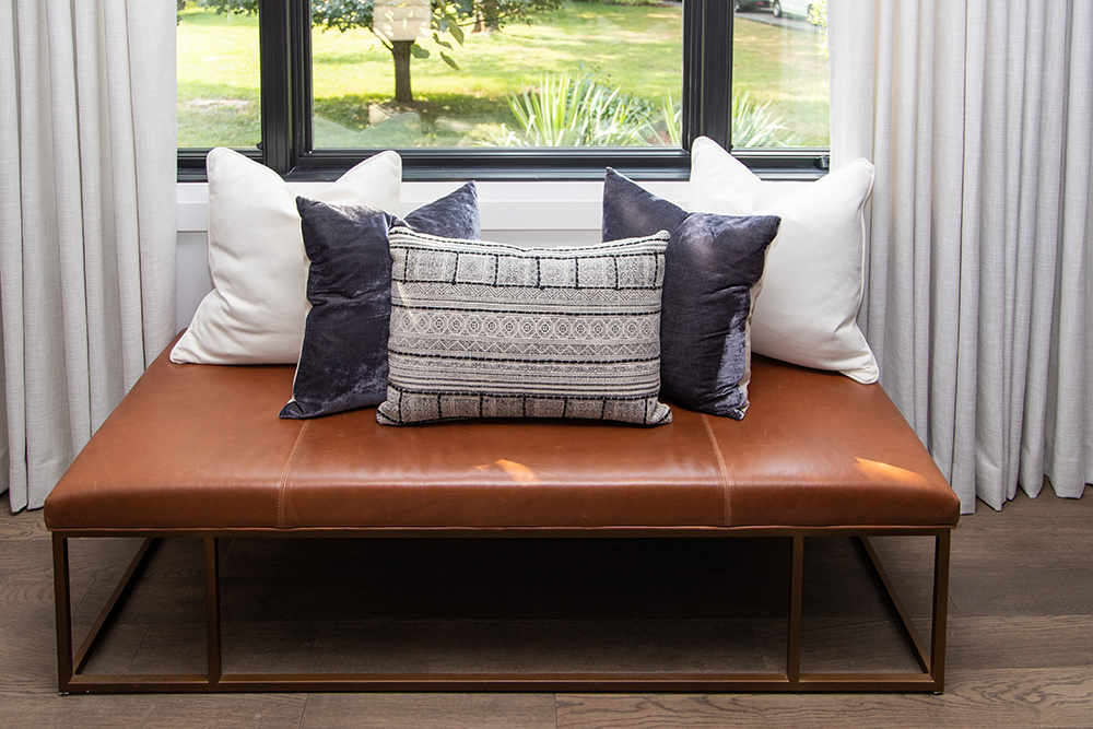 A chic leather ottoman bench sent stacked with five throw pillows in white, purple and black.
