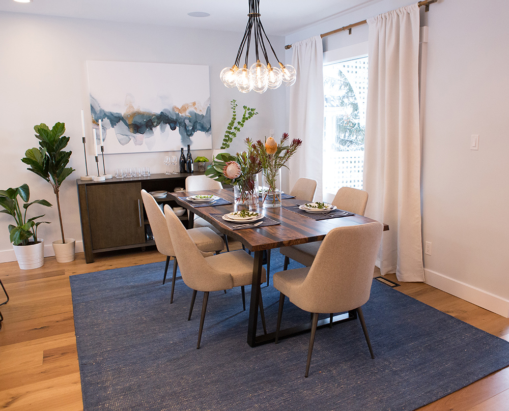 Gorgeous formal dining room featured on The Property Brothers on HGTV with six sand coloured unposted chairs, a large live edge wood table, two glass vases filled with usual flowers, a dark wood sideboard, a large watercolour print on the wall, and blue woven rug on the floor