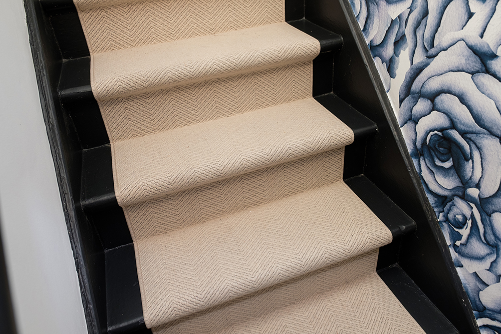 A cream coloured runner goes up black stairs against a wall of blue rose wallpaper