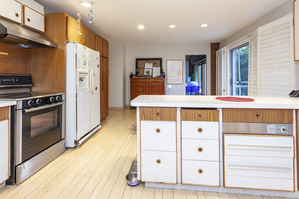 1980s kitchen with white laminate cabinets, black oven, white fridge and worn out blonde wood flooring