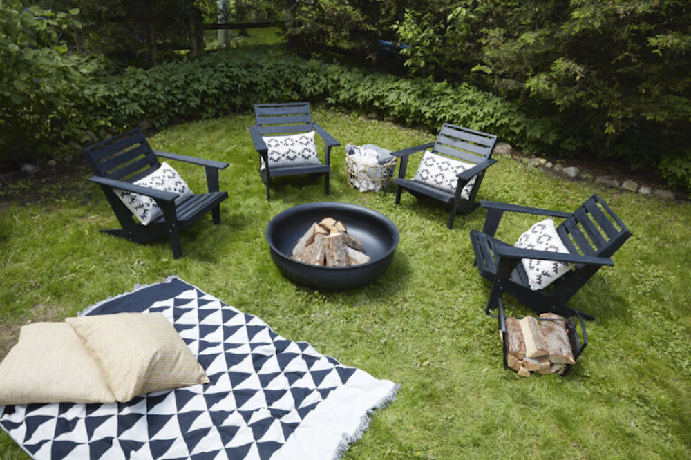 A black fire pit filled with logs surrounded by four black lawn chairs and a black and white blanket spread on the grass with two pillows as featured on Home to Win on HGTV