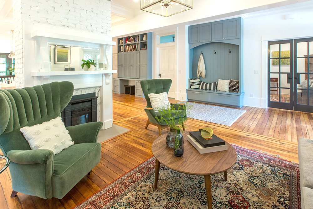 Beautiful living room featuring stained hardwood floors, two green velvet wing backed chairs, a red woven rug, a round wooden coffee table, a white brick fireplace, a blue coat nook, and white walls as featured on Property Brothers on HGTV Canada