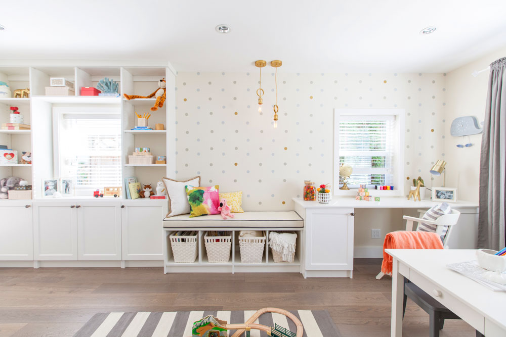 Playroom designed by Jillian Harris on HGTV Canada with built-in wraparound window cubbies with a cushioned seat, quirky gold knotted pendant lamps, white walls, polka dot wallpaper, dark wood floors, and a grey and white striped rug