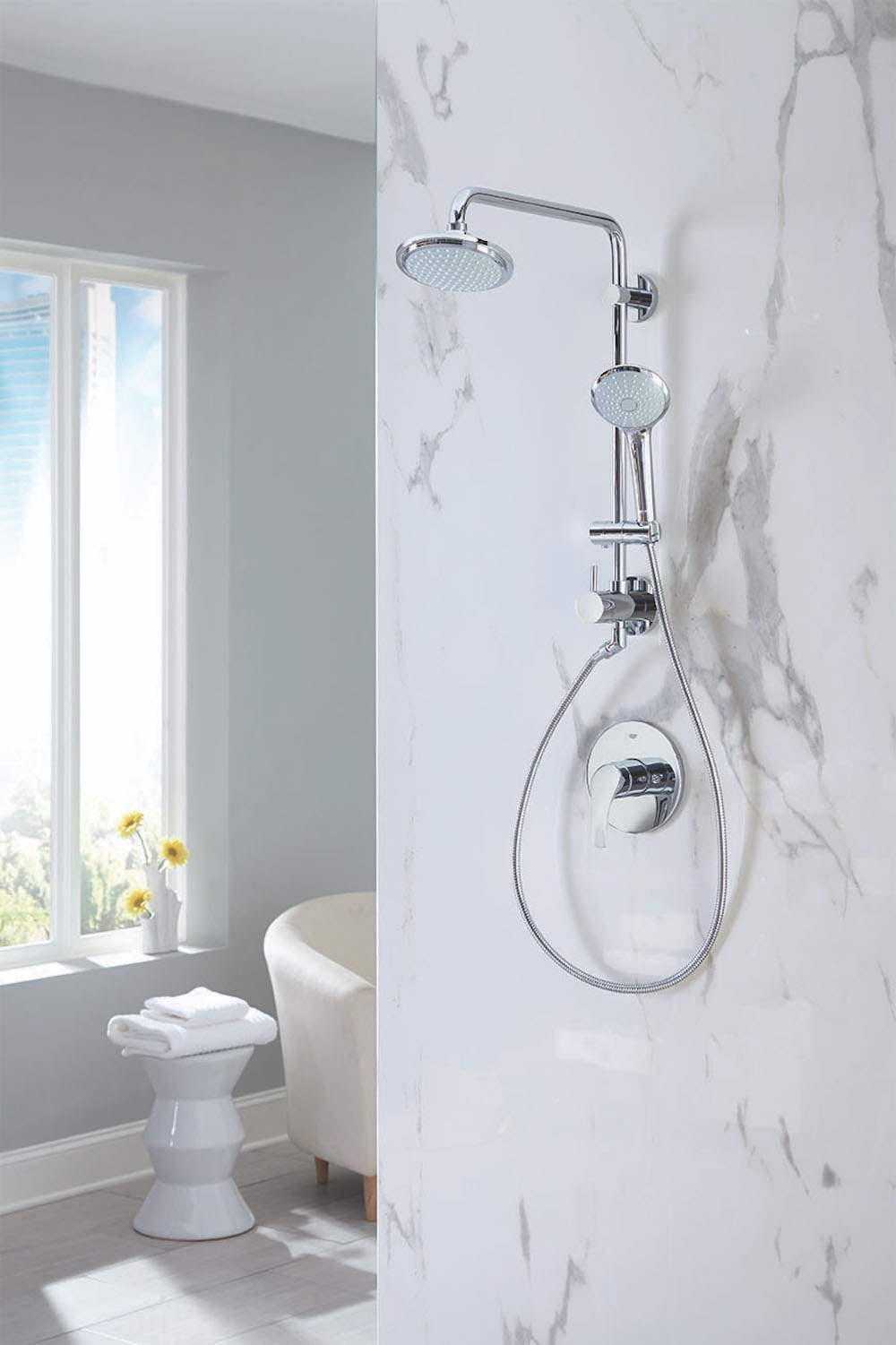 Modern shower with large grey and white marble walls and a GROHE Retrofit 260 Shower System from Splashes Bath & Kitchen, grey tiled floors, a white stool with white towels folded onto, a window with a white vase with yellow flowers, a grey blue wall, and the corner of a white armchair