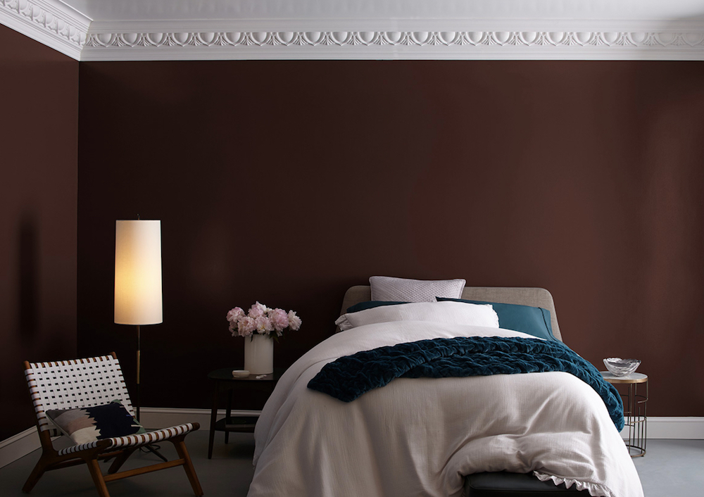 A simple bedroom with an upholstered bed dressed with white and teal bedding, a wooden mid century modern chair with white cloth slaps, crown moulding on the ceiling painted in BEHR Ultra Pure White 1850, and walls painted in a deep red with BEHR Oxblood PPU2-20