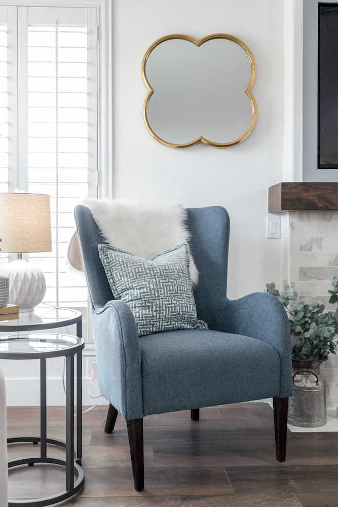 Cool blue armchair with throw pillow and faux sheepskin rug, clover shaped gold mirror, dark vinyl floors and set of two round glass side tables