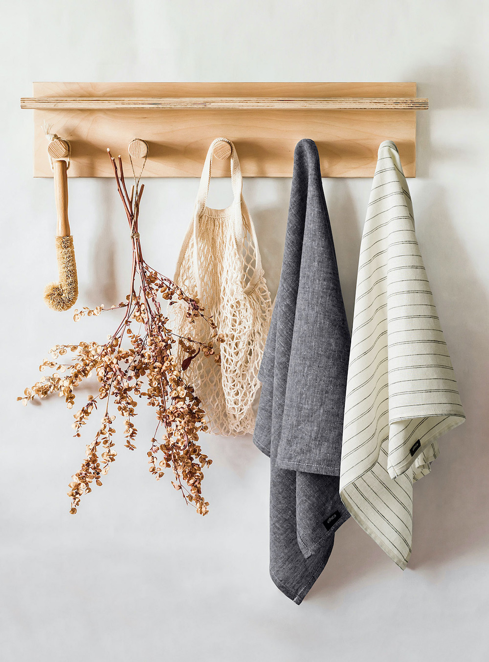 A set of refined yet casual striped hemp and organic cotton cream and grey dish towels hanging from a wooden wall rack along with a net bag, dried flowers and sisal scrub brush