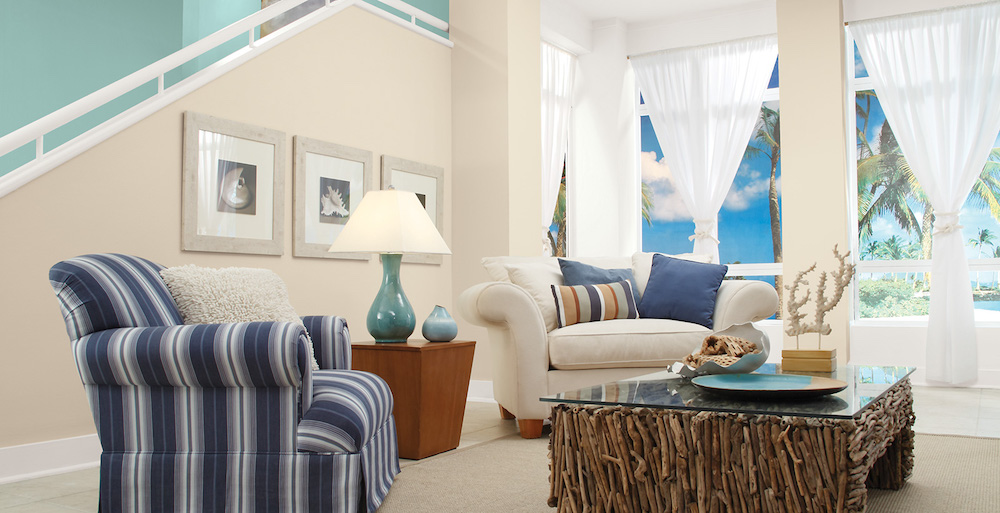 A chic beach-themed living room with a driftwood coffee table, three shell photos on the wall, sheer white drapes, a white couch and blue-striped armchair covered in blue throw pillows, and walls painted in BEHR Roman Plaster PPU7-10, Palais White GR-W15 and Opal Silk PPU12-08