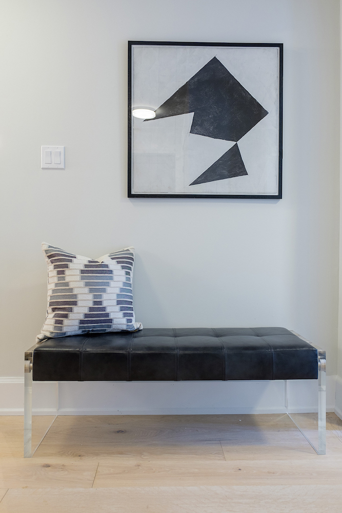 Simple foyer with light wood floors, a black leather and Lucite bench, a framed black and white art, and a white, grey and blue throw pillow