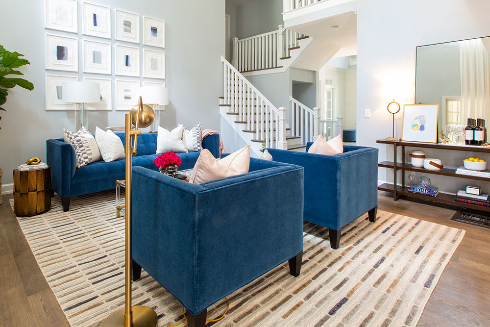 Chic living room with two blue velvet armchairs, a tufted blue velvet couch, a gallery wall of white framed art, a gold floor lamp, and set of stairs and landings