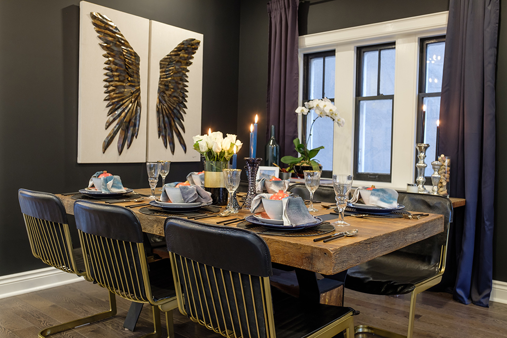 A thick wood dining table surrounded by black leather and brass dining chairs with a piece of winged art on the wall