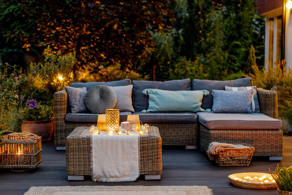 Beautiful wooden deck lit up at night with a large wicker couch and coffee table covered in lit candles surrounded by mature plants