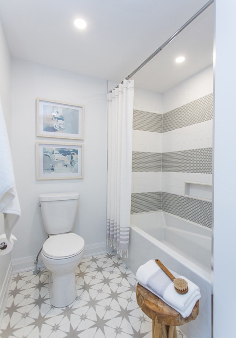 Small white bathroom with large grey star statement tile on the floor, a bathtub shower combo with small grey and white penny tiles, and two framed prints above a white toilet