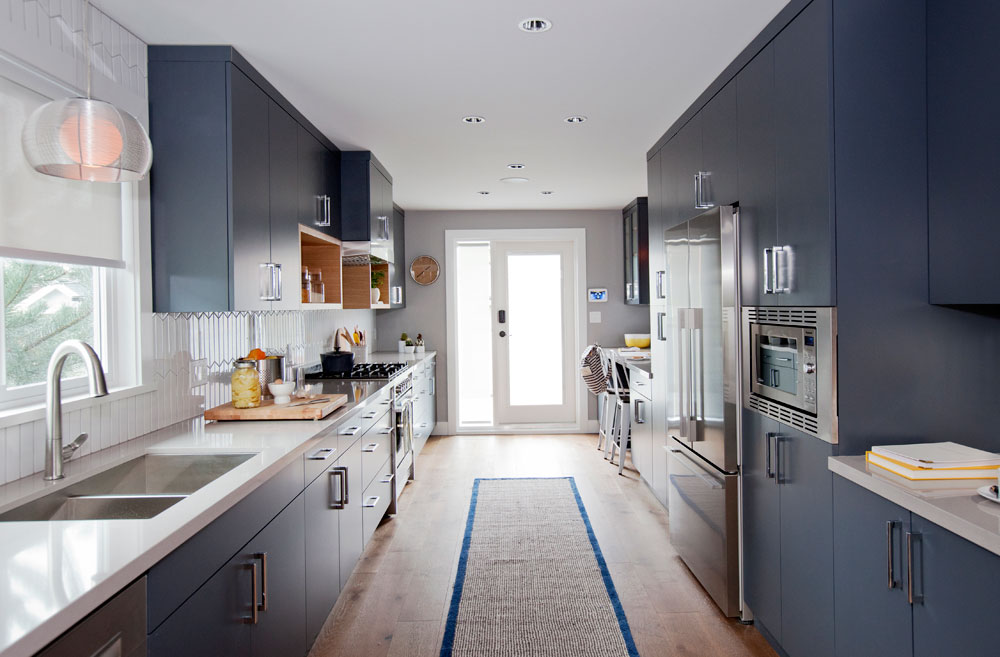 Narrow kitchen with sleek blue cabinetry, modern white backsplash and light wood flooring.