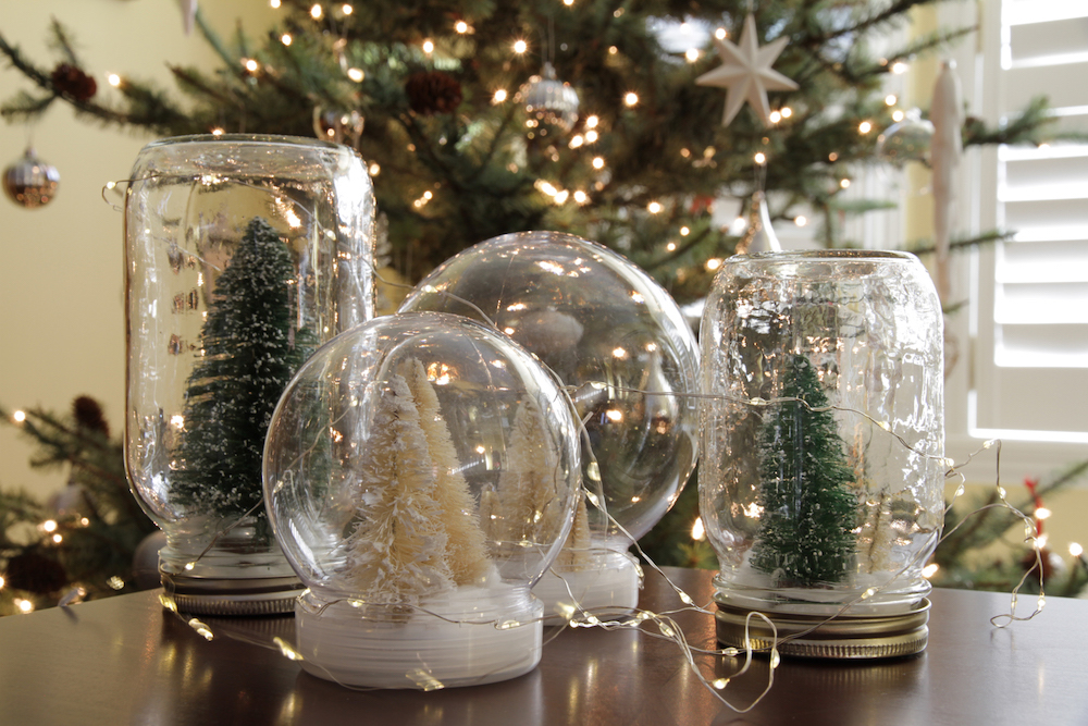 Four pretty DIY snow globes sit on a table in front of a Christmas tree