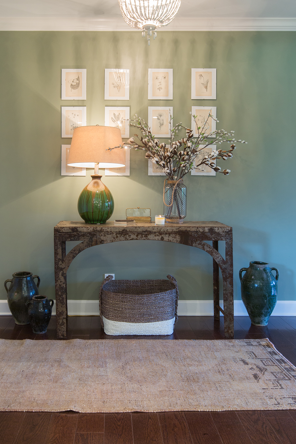 Vintage entry table topped with a large green ceramic lamp, a vase filled with cotton sprigs, three large ceramic vessels on the ground, a large woven basket on dark wood floors, a vintage rug, and a green wall with 12 framed vintage botanical prints hung on it