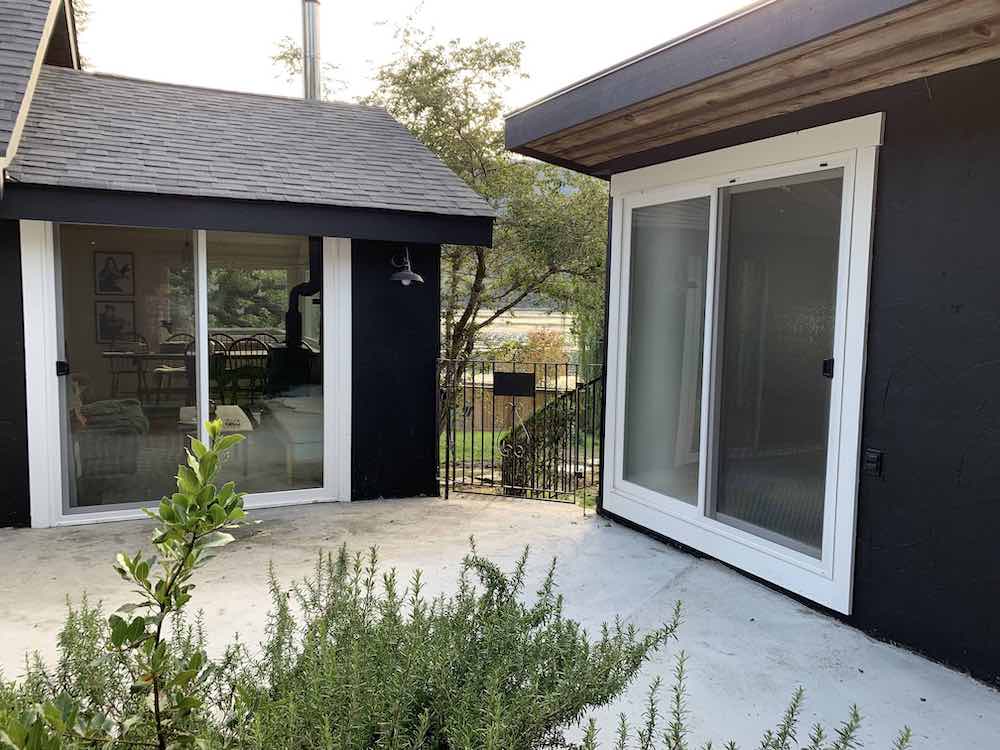 Exterior of a cottage courtyard with a large rosemary bush, greenery growing in behind it, and walls painted black with BEHR Black Smoke PPH-34 and window and door trim painted with BEHR Ultra Pure White 1850