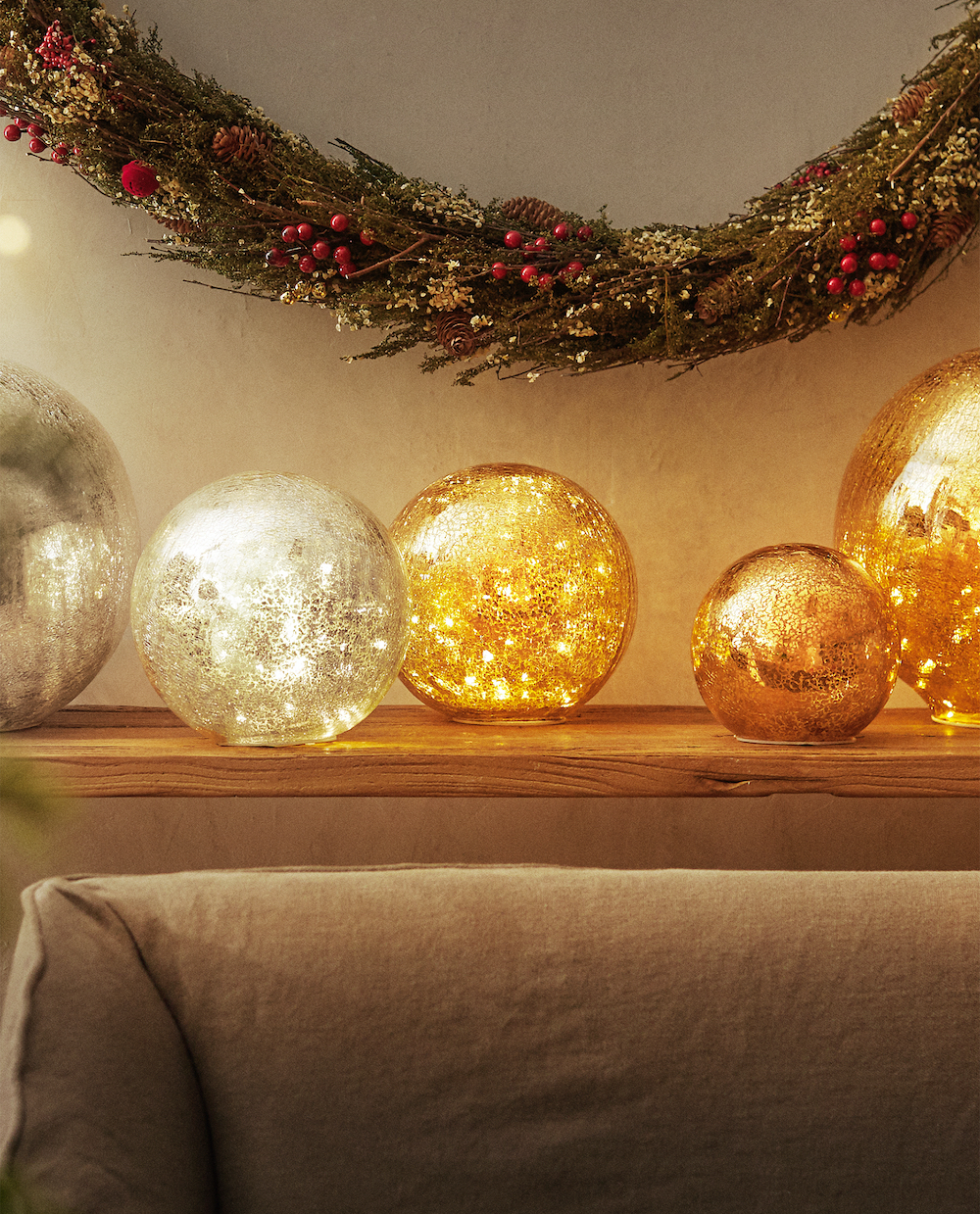 Zara Home interior crackled glass light Christmas ornaments on a wooden shelf above a beige couch back and a long Christmas garland hanging on the wall