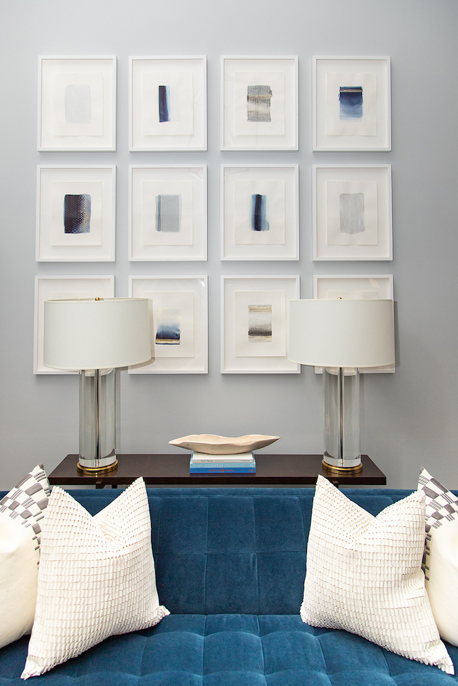 Chic living room with a tufted blue velvet couch, a side table with two Lucite lamps, and a wall of 12 white framed watercolour paintings