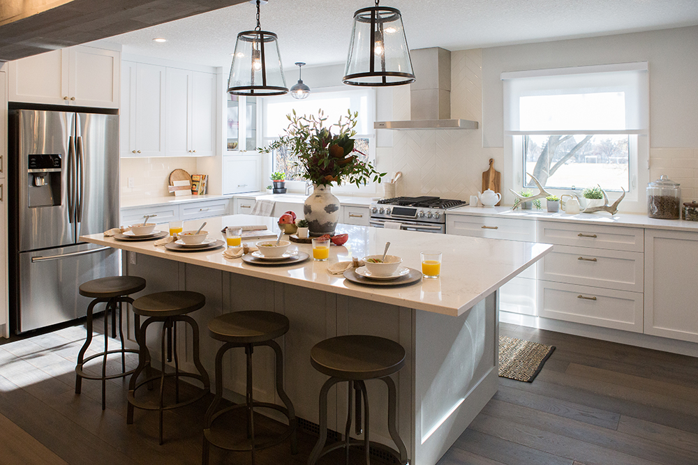 Bright and modern kitchen featured on The Property Brothers on HGTV with a white island set for breakfast, with four stools, two large glass and iron pendant lights, and a stainless-steel fridge