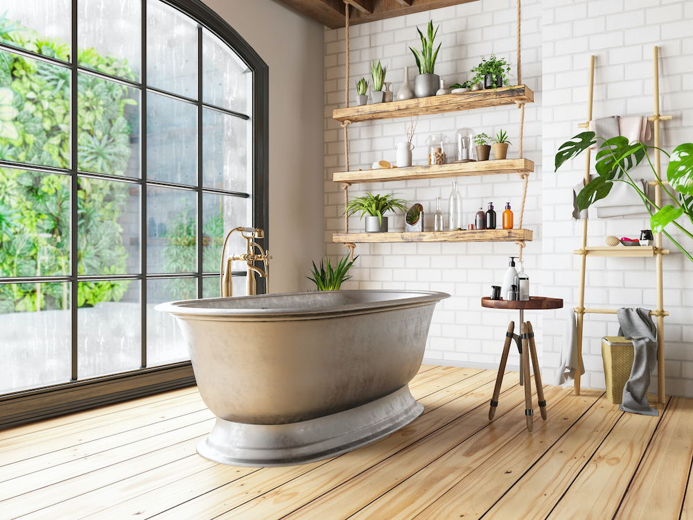 Beautiful bathroom with a large steel pedestal bathtub sitting in front of a large black framed window, wide plank wooden floors, a stool topped with soaps and lotions, a bamboo ladder leaning against the wall with towels hanging on it, a large plant, and a set of three shelves hanging from the ceiling with role filled with potted plants, bottles and objets d’art