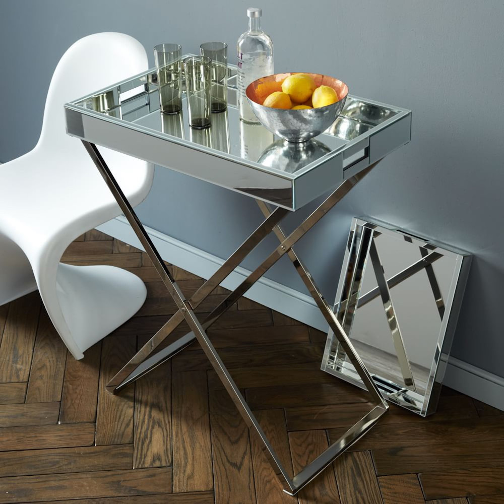 A white modern chair and a mirrored tray on a silver stand holds a silver bowl of lemons, a bottle of vodka and three glasses against a grey wall