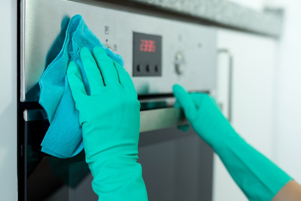 Close up of hands in green gloves cleaning the outside of a stove.