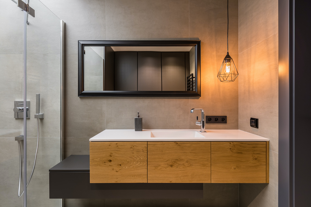 Industrial design bathroom with large grey concrete effect tiling on the walls, a floating wood vanity with a white countertop and sink with silver hardware, a rectangular black framed mirror on the wall about it along with a black cage pendant lamp, and a glass wall shower to the left