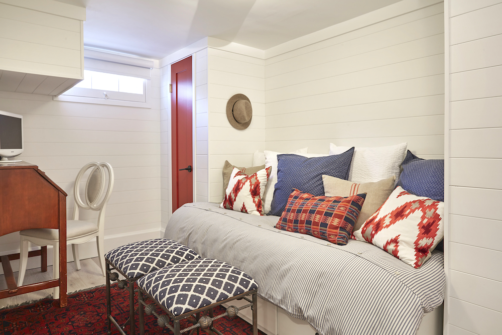 Buyers Bootcamp detached bungalow basement bedroom with white wood panel siding, a red door and a bed covered in red, blue and white pillows