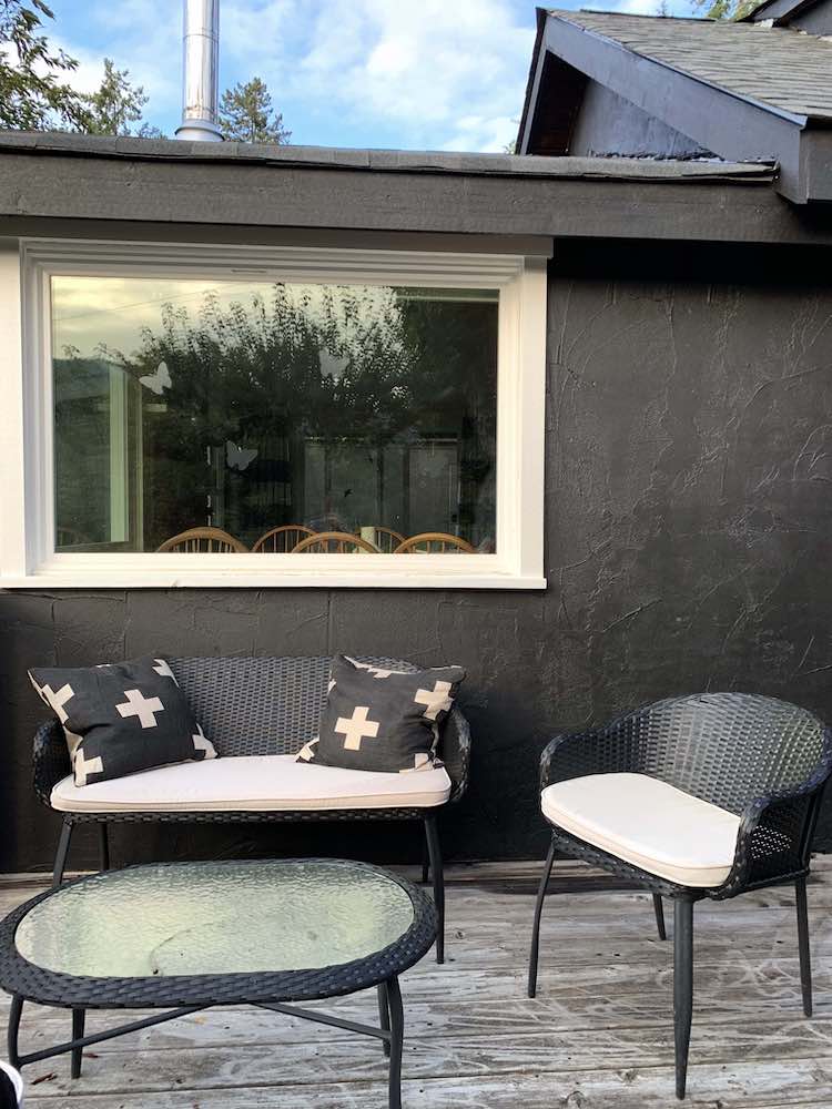 Exterior of a cottage with a bench with two cushions, a chair, a glass-topped table, and walls painted black with BEHR Black Smoke PPH-34 and window painted with BEHR Ultra Pure White 1850