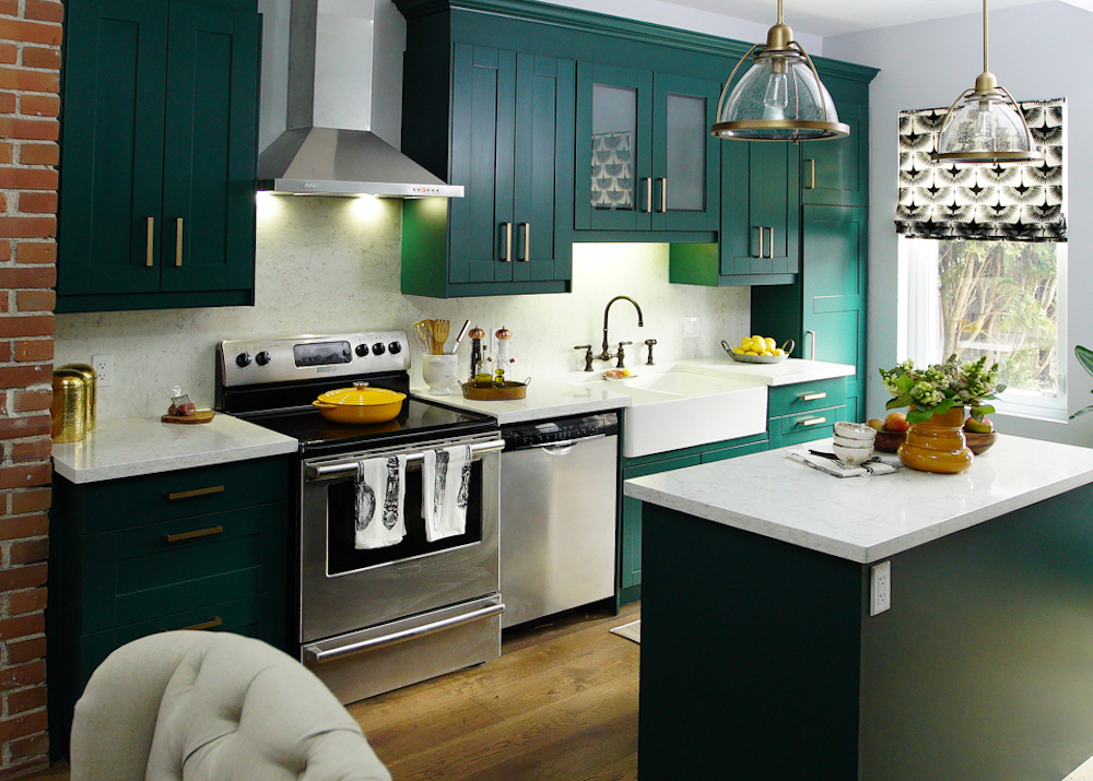 Beautiful newly renovated kitchen with emerald green cabinets, white and grey quartz countertops, stainless steel hood fan, and a small kitchen island with two glass and brass pendants lights hanging above it as featured on HGTV’s Save My Reno