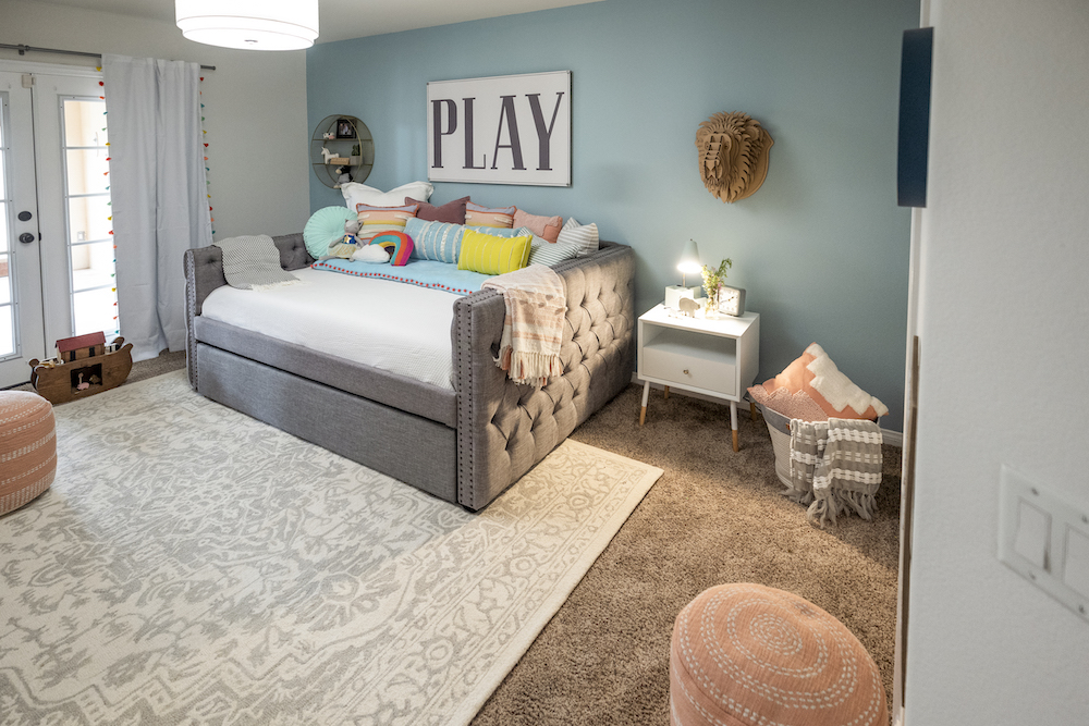 A children’s play room with large grey tufted day bed, a blue wall, large white and grey area rug and a sign on the wall that says “Play”