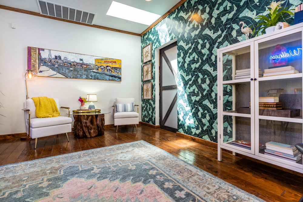 An open office space with palm leaf wallpaper, two white armchairs, a driftwood coffee table, large colour area rug and a long colourful wall painting